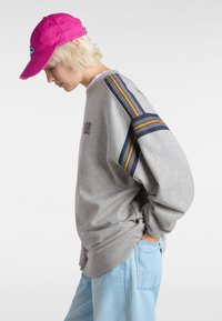 Grijze sweatshirt met geribbelde manchetten, met marineblauwe en mosterdgele strepen op de mouwen. Draagt het met lichtblauwe broek en een felroze pet.