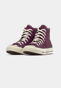 Burgundi spalvos aukštakulniai drobės sportbačiai su baltais raišteliais ir guminiu padu, esančiais ant šviesiai pilko fono.