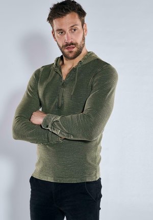 Grüne Zip-Hoodie aus strukturiertem Stoff mit Kapuze und Zugband sowie umgeschlagenen Bündchen, kombiniert mit schwarzen Hosen. Neutraler Hintergrund.