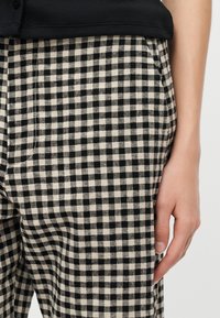 Δεξί χέρι να ακουμπά δίπλα σε ασπρόμαυρο παντελόνι με σχέδιο καρό, με ορατή πλαϊνή τσέπη και μέρος από μαύρο μπλουζάκι.