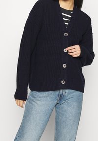 Námořnický modrý pletený cardigan s výstřihem do V, hrubou strukturou, čtyřmi předními knoflíky a manžetami se žebrováním, kombinovaný se světlými modrými džíny.