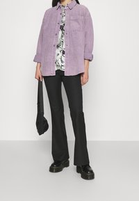 Ljust lila kordflöjel knapp-up skjorta över en mönstrad vit topp, tillsammans med svarta utsvängda byxor och svarta kraftiga skor, samt en svart väska.