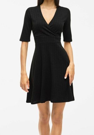 Kobieta nosząca dopasowaną czarną prążkowaną dzianinową sukienkę z rękawami do łokci i dekoltem w serek, stojąca na jednolitym tle.