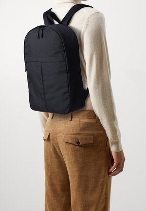 Personne portant un col roulé beige et un pantalon marron avec un sac à dos noir minimaliste dans le dos, devant un fond uni.