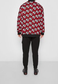 Chaqueta bomber negra con patrón de texto "HUGO" en rojo y blanco, combinada con pantalones negros ajustados y zapatos negros con detalles de logo en rojo.