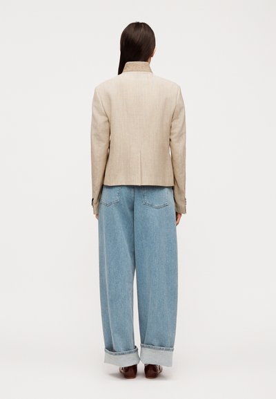Γυναίκα με μακριά σκούρα μαλλιά που φοράει μπεζ σακάκι, μπλε τζιν παντελόνι με φαρδιά παντελόνια που έχουν γυρισμένα ραφή και καφέ παπούτσια, στέκεται γυρισμένη με την πλάτη σε λευκό φόντο.