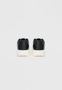 Mustat lenkkitossut, joissa on teksturoitu päällinen ja sileä valkoinen pohja. Niissä on kohotettu geometristen kuvioiden malli kannassa ja sivuilla lisäyksinä.