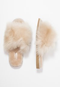 Chaussons en fausse fourrure beige avec un matériau doux et moelleux, design à bout ouvert, semelle plate et texture semblable à du daim sur la semelle intérieure et la semelle extérieure.
