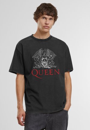 Jeune homme portant un t-shirt ample noir du groupe Queen avec un blason blanc et un texte rouge, associé à un jean foncé, debout devant un fond uni.