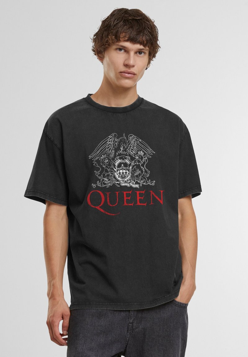 Jeune homme portant un t-shirt ample noir du groupe Queen avec un blason blanc et un texte rouge, associé à un jean foncé, debout devant un fond uni.