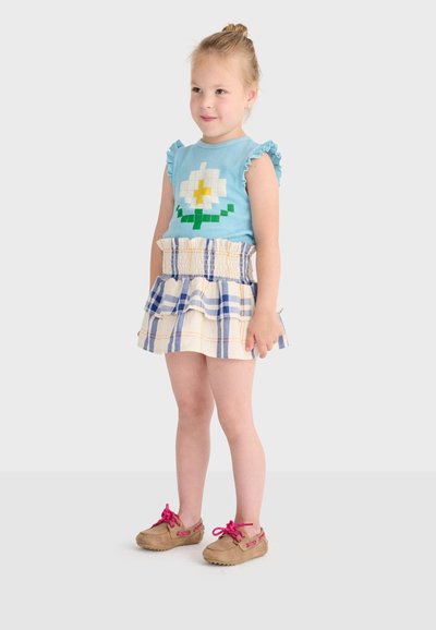 Ung pige stående iført en blå ærmeløs bluse med blomsterdesign, en flæset ternet nederdel og brune sko med lyserøde snørebånd.