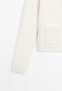 Cardigan color crema con superficie testurizzata, maniche lunghe e una tasca applicata. Rifinito con orlo e polsini a coste.