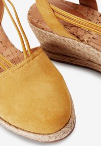 Sandales à talons compensés en daim jaune avec une semelle en liège et en jute, présentant un bout arrondi et des détails de sangle avec des accents jaunes.