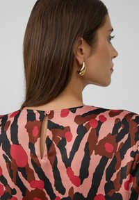 Rosa Stoffoberteil mit schwarzen und roten abstrakten Mustern, featuring einem Schlüsselrücken und kleinen runden Knopfdetails. Große goldene Ohrringe.