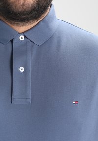 Pikétröja i ljusblått bomull med en strukturfinish, med en knappslå i tre knappar och en liten logotyp på vänster bröst.