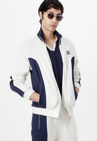 Veste de sport blanche et marine avec col montant et fermeture éclaire, associée à un haut marine. Le modèle porte des lunettes de soleil, affichant une coupe décontractée.