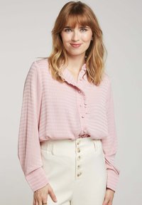Chemisier rose clair à boutons avec un col à volants, en tissu texturé avec des rayures verticales, associé à un pantalon blanc taille haute avec des boutons.
