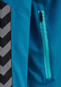 Blauwe sportjack met een zwart-wit chevronpatroon op het paneel. Inclusief een ritssysteem met een turquoise ritsdetail.