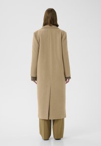Cappotto di lana beige con silhouette dritta, maniche lunghe e spacco centrale sul retro. Indossato sopra polsini a coste verde oliva e pantaloni marroni.