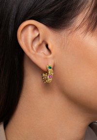 Orecchini a cerchio dorati con motivi floreali, adornati con gemme verdi, rosa e viola, esposti su un orecchio.