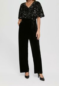 Svart paljettopp med V-ringning och kapärmar, matchad med svart flanellbyxor med hög midja och vida ben. Svarta högklackade skor ingår.