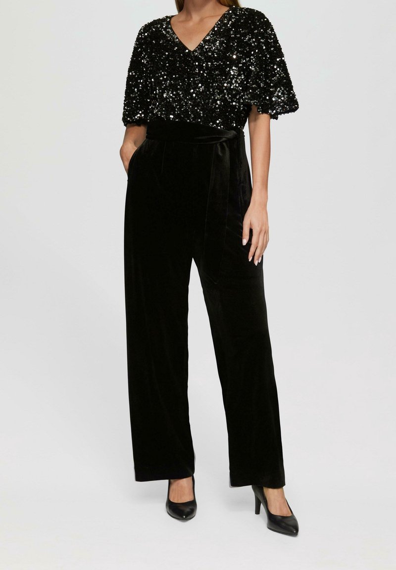 Svart paljettopp med V-ringning och kapärmar, matchad med svart flanellbyxor med hög midja och vida ben. Svarta högklackade skor ingår.