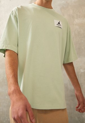 Mężczyzna ubrany w jasnokołową, oversize'ową koszulkę z małą białą naszywką z czarnym logo w kształcie sylwetki na piersi, stojący przy szarej ścianie.