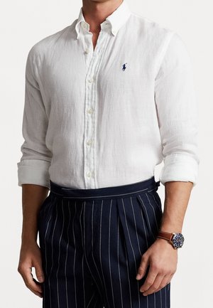 Homme portant une chemise en lin blanche boutonnée aux manches retroussées, un pantalon bleu marine à fines rayures et une montre à bracelet en cuir marron au poignet gauche.