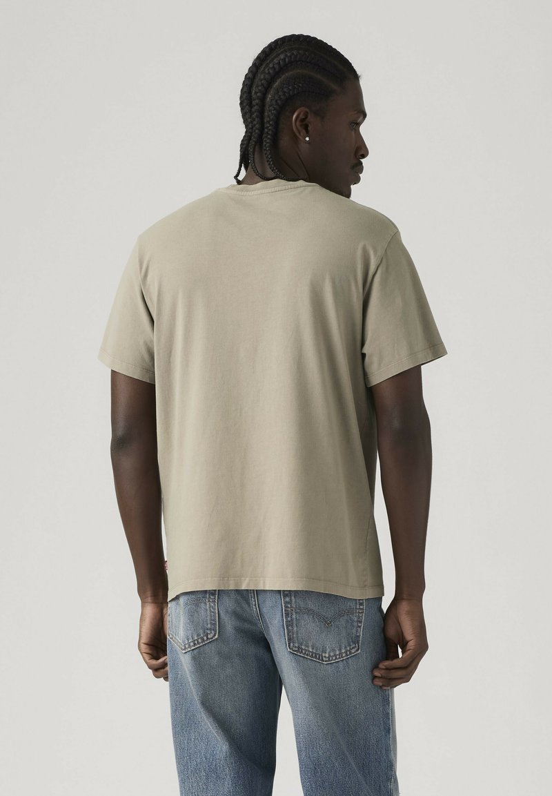 Homme aux cheveux tressés de dos, portant un t-shirt beige à manches courtes et un jean bleu, sur un fond clair uni.