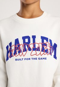 Sudadera blanca con gran texto azul "HARDLYM", superposición en estilo graffiti rojo y texto azul más pequeño "BUILT FOR THE GAME" debajo del texto principal.