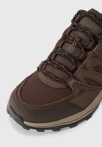 Brown hiking shoe featuring mesh and suede materials, black laces with orange accents, textured sole, and reinforced toe cap.