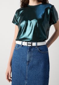 Skinande smaragdgrön kortärmad skjorta i reflekterande tyg, matchad med en högmidjad blå denimkjol och ett vitt bälte.