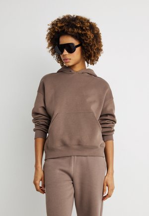 Kvinne med krøllete hår iført mørke solbriller, en brun hettegenser og matchende bukser, stående foran en enkel lys bakgrunn.