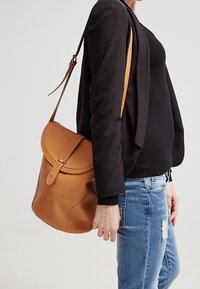 Kožený batoh v barvě tan s kulatým tvarem, klopovým zavíráním a nastavitelným popruhem. Nosený přes rameno, v kombinaci s černým topem a denimovými kalhotami.