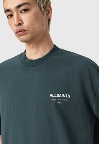 Gros plan sur une personne portant un T-shirt vert foncé avec "ALLSAINTS" et des caractères japonais imprimés en blanc sur la poitrine.