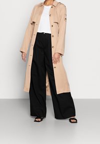 Beige lång trenchcoat med snittkrage, framknappar, två sidofickor, bärs över en vit topp och svarta vida byxor. Svarta sandaler.