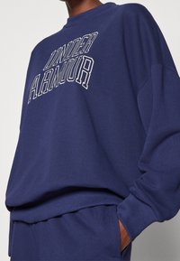 Sudadera azul marino de manga larga, con puños acanalados y cuello redondo. Presenta el logotipo "UNDER ARMOUR" en letras blancas en la parte frontal.