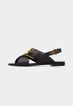 Black flat sandal with wide crisscross straps and two gold buckle closures, one large on the front and a smaller one on the heel strap.