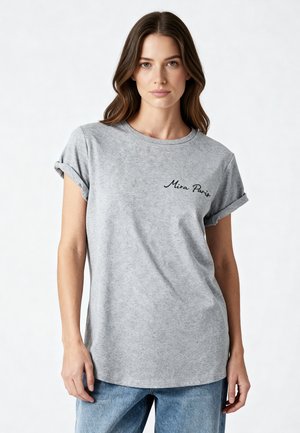 Femme aux longs cheveux bruns portant un t-shirt gris à manches retroussées avec le texte « Mira Paris » sur la poitrine et un jean bleu sur un fond uni.