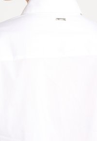 Vitskjorta med krage som har en fräsch textur, slät tygkvalitet och en liten metallisk logotagg vid nackområdet.