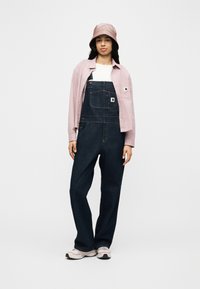 Γυναίκα με σκούρα τζιν φορμά, λευκό πουκάμισο, ανοιχτό ροζ υφασμάτινο μπουφάν, ροζ καπέλο τύπου μπούκετ που ταιριάζει και λευκά αθλητικά παπούτσια, στέκεται μπροστά σε απλό φόντο.