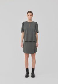 Graues, kurzärmeliges Kleid mit Schlüsselloch-Ausschnitt und geschichtetem Design. Glatter Stoff, knielang, kombiniert mit schwarzen Knöchelschuhen.