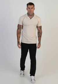 Beige polo shirt with a collar, fitted design, paired with black pants. Model wearing white sneakers, standing on a light background.