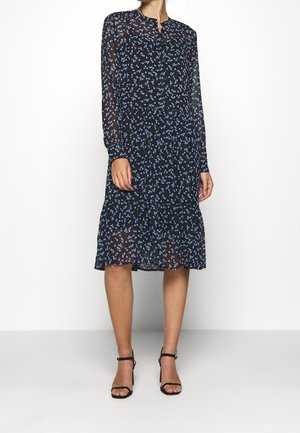 En kvinde iført en sort midikjole med lange ærmer med små blå blomsterprint og sorte høje hælsandaler, står mod en hvid baggrund.