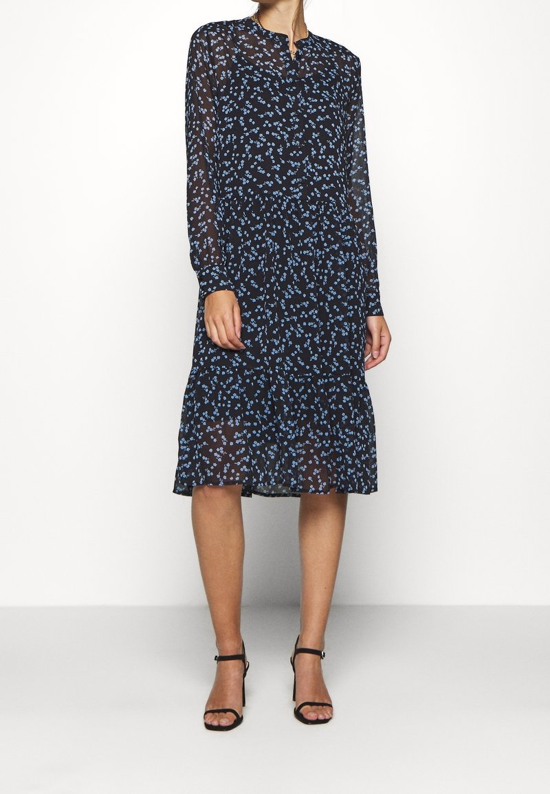 Femme portant une robe midi noire à manches longues avec un petit imprimé floral bleu et des sandales noires à talons hauts, debout sur un fond blanc.