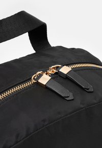 Närbild av en svart väska med dragkedja med guldtenn och svarta läderdragflikar, samt ett svart vävt tyghandtag ovanför dragkedjan.