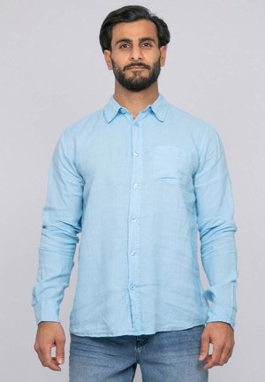 Homme barbu portant une chemise bleu clair boutonnée et un jean bleu, debout devant un fond gris clair, regardant vers l'avant.