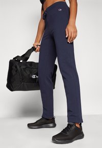 Marina träningsbyxor med dragsko i midjan, kombinerat med svarta träningsskor och en svart duffelväska med en diskret logotyp.