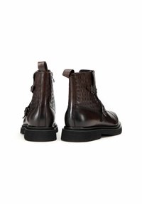 Botas de tobillo de cuero marrón con textura de cocodrilo. Incluyen un cierre de hebilla, una lengüeta para tirar y una suela de goma negra gruesa.