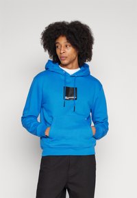 Blauwe hoodie van zacht materiaal, met een voorzak, verstelbare trekkoorden en een zwart logo-patch op de borst. Zwarte broek gedragen.